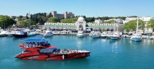 Rhodes Sea Lines boat with passengers docked in a turquoise harbor, with a historic fortress on a tree-lined hill in the background and a clear blue sky.